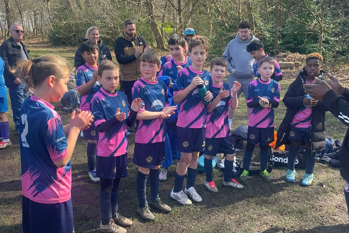 Applauding at the end of the match in the pink and navy away kit, on a woodland pitch