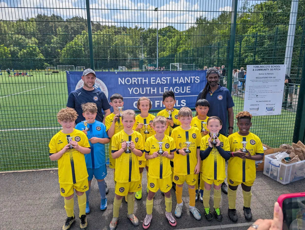 The squad and their coaches with trophies under the North East Hants Youth League Cup Finals banner