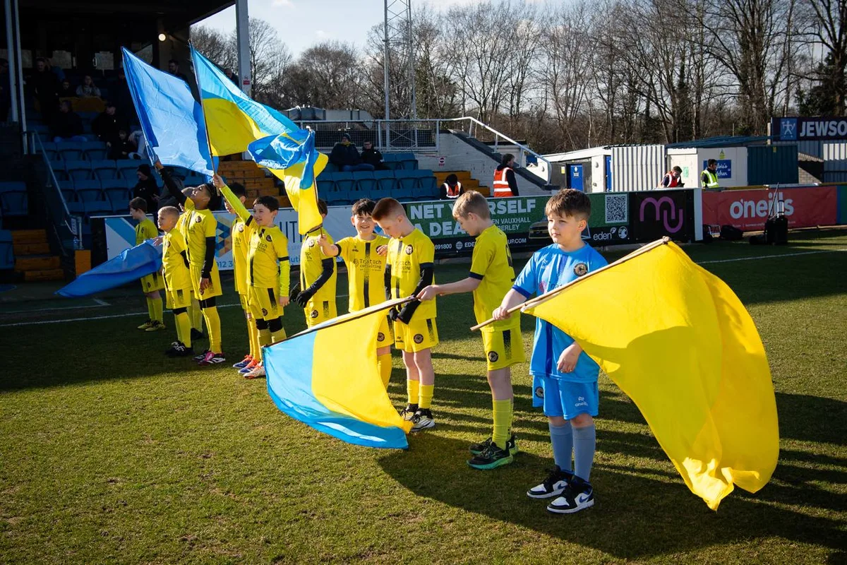 Young mascots lined up across the pitch with the club's blue and yellow flags before a Farnborough FC first-team match