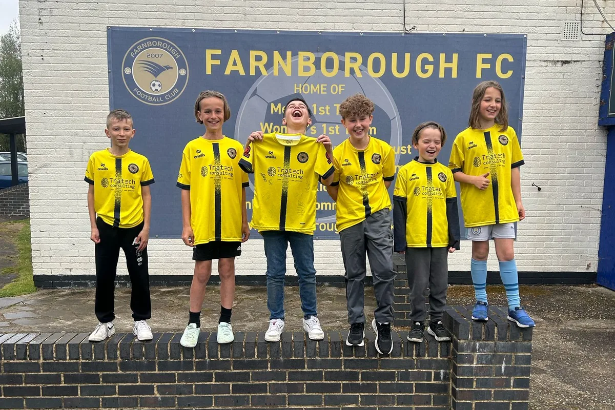Six players lined up along the wall under the Farnborough FC sign, holding a signed shirt between them