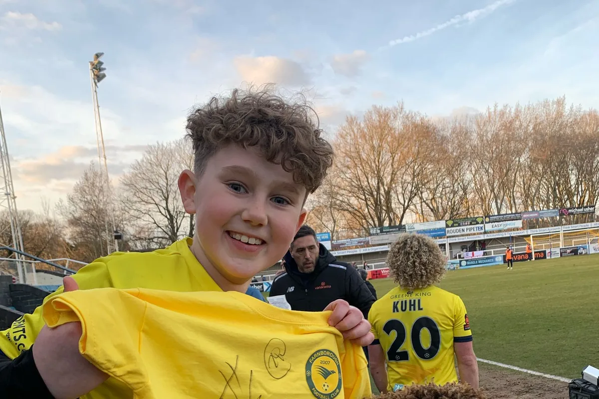 A young supporter holding up a signed Farnborough FC shirt at the side of the pitch