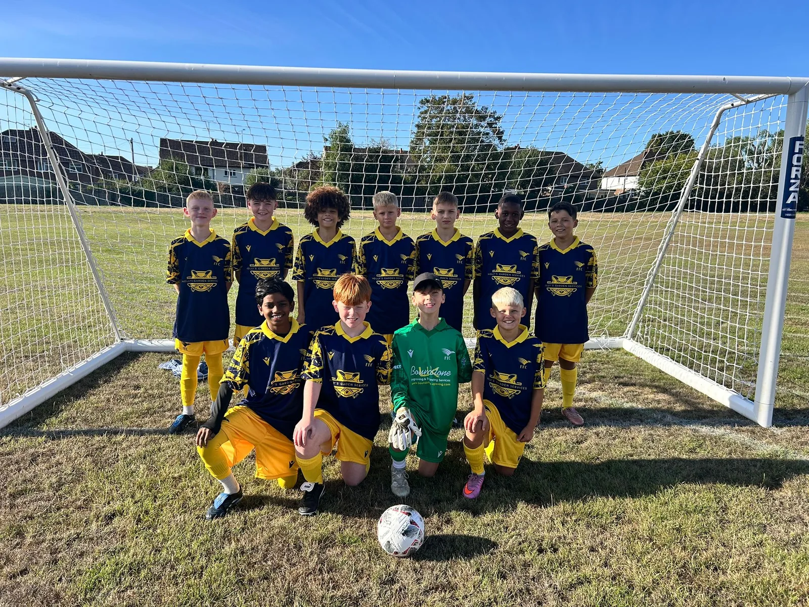 The Under 12 Yellows in their navy and yellow away kit, seven standing and four kneeling behind the matchball