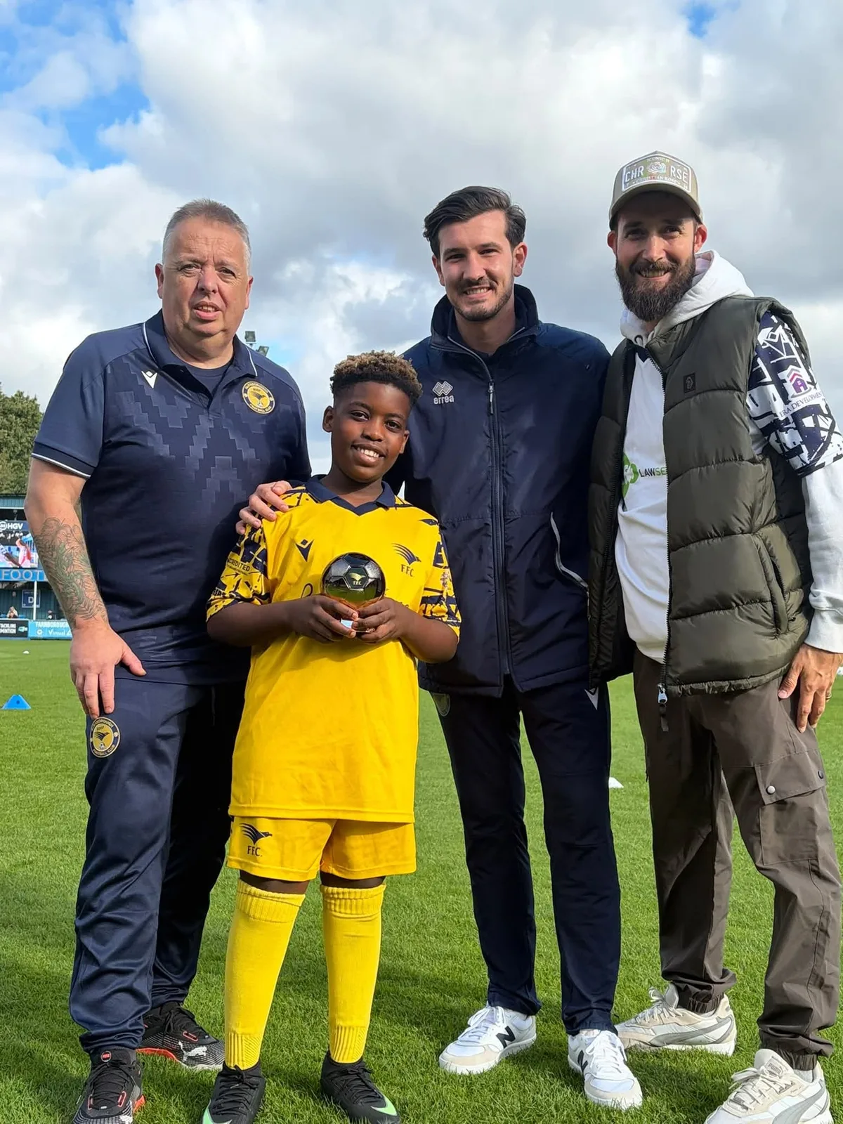 A beaming young player in the yellow club kit holds a gold football trophy on the stadium pitch, with three adults from the club alongside