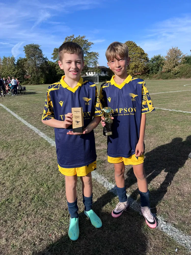 Two team-mates in the navy away kit, side by side on the pitch, each holding a trophy