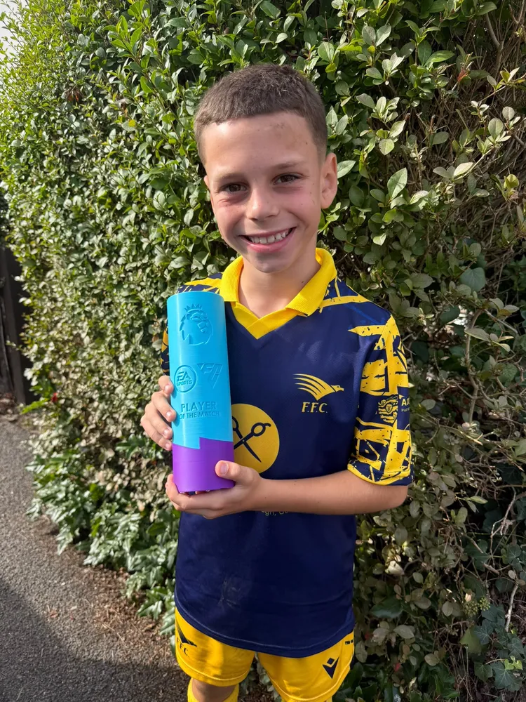Grinning by a hedge in the navy away shirt, with a tall turquoise and purple trophy