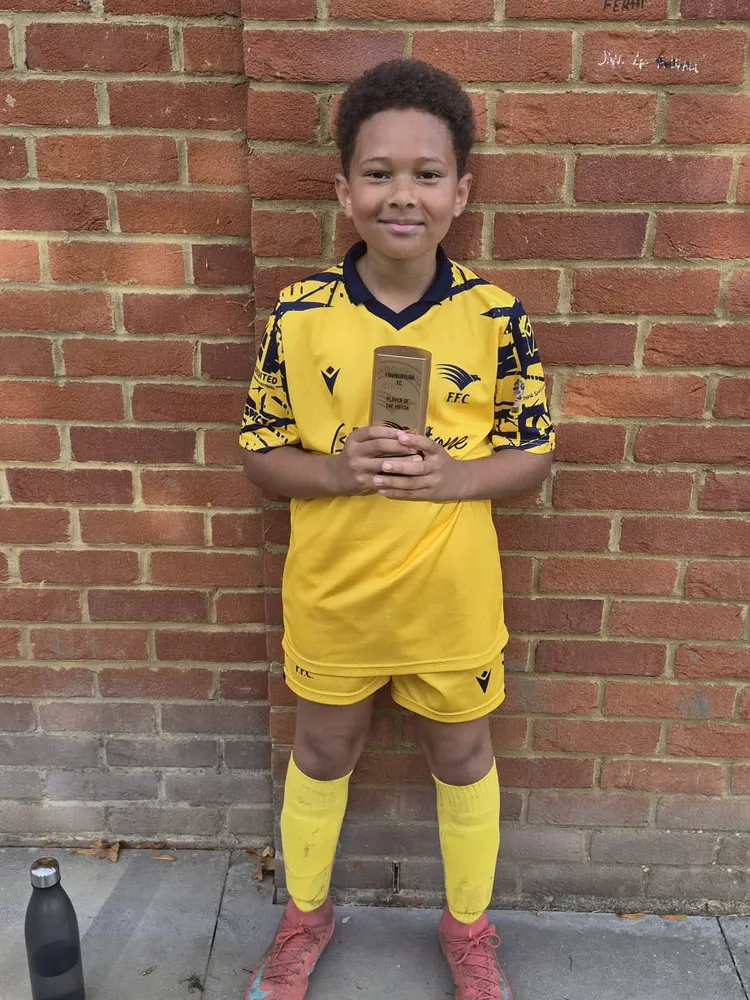 Against a brick wall in red boots, a small gold trophy in both hands