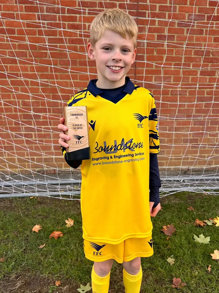 In front of a brick wall and goal net, a gold Player of the Match trophy held up beside a big smile
