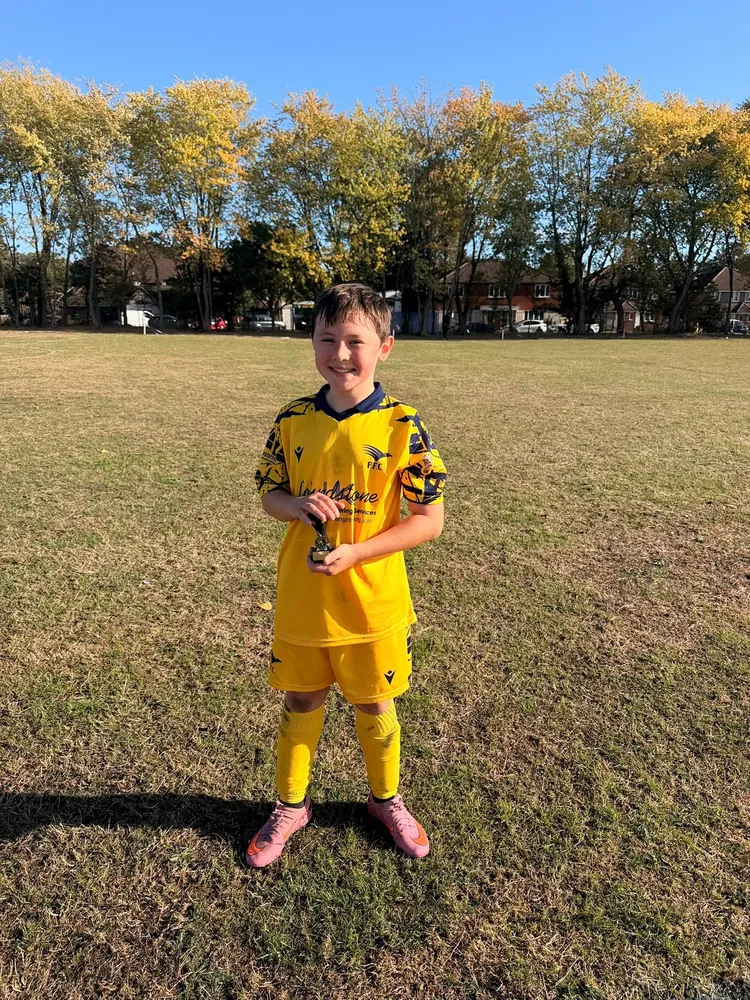 Pink boots and a small gold trophy on a wide, sunlit pitch lined with autumn trees