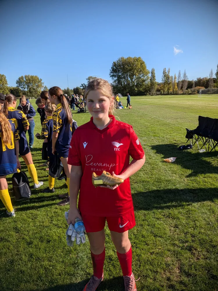 Keeper's gloves and a water bottle in one hand, a golden boot trophy in the other, with teammates in navy and yellow chatting behind