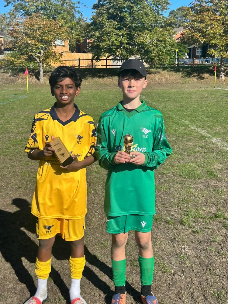 Two teammates side by side on a sunny pitch, one in yellow and one in the keeper's green, each holding a trophy
