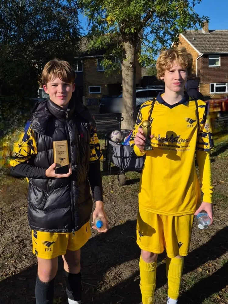 One in a black puffer jacket and one still in yellow kit, two trophies held up under a big tree