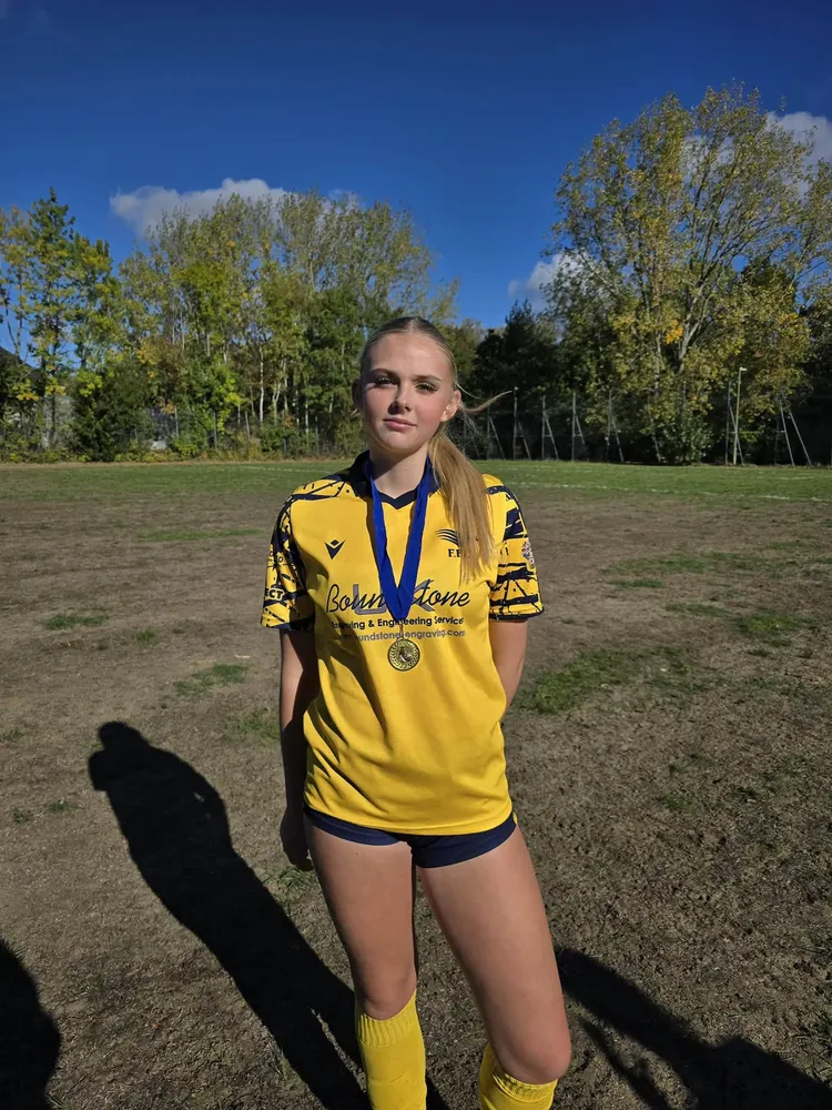 A gold medal on a blue ribbon and a calm, steady gaze, with long afternoon shadows stretched across a bare patch of pitch