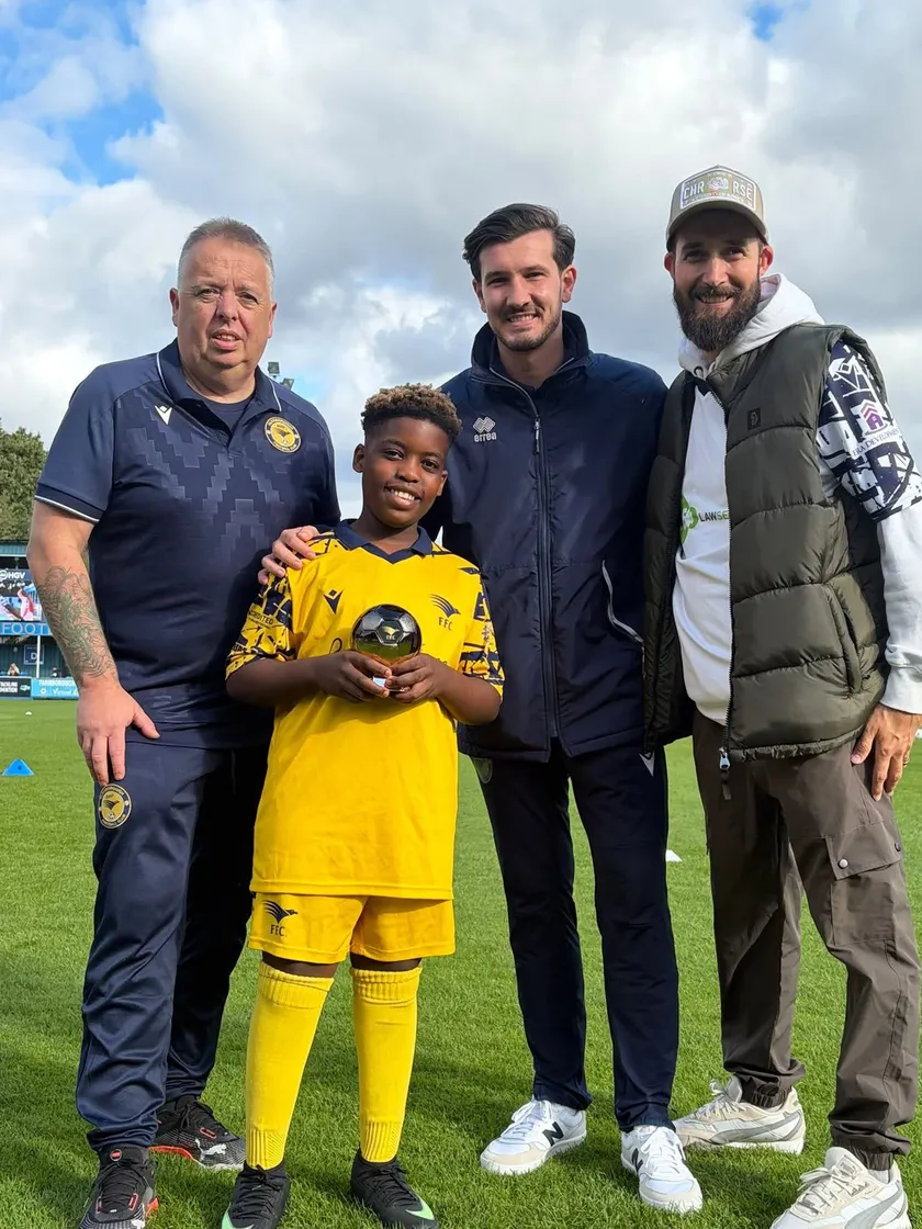 A beaming young player in the yellow club kit holds a gold football trophy on the stadium pitch, with three adults from the club alongside