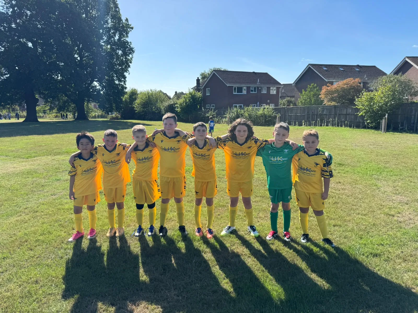 The Under 10 Yellows squad lined up arm in arm across the grass on a bright morning, long shadows stretching towards the camera