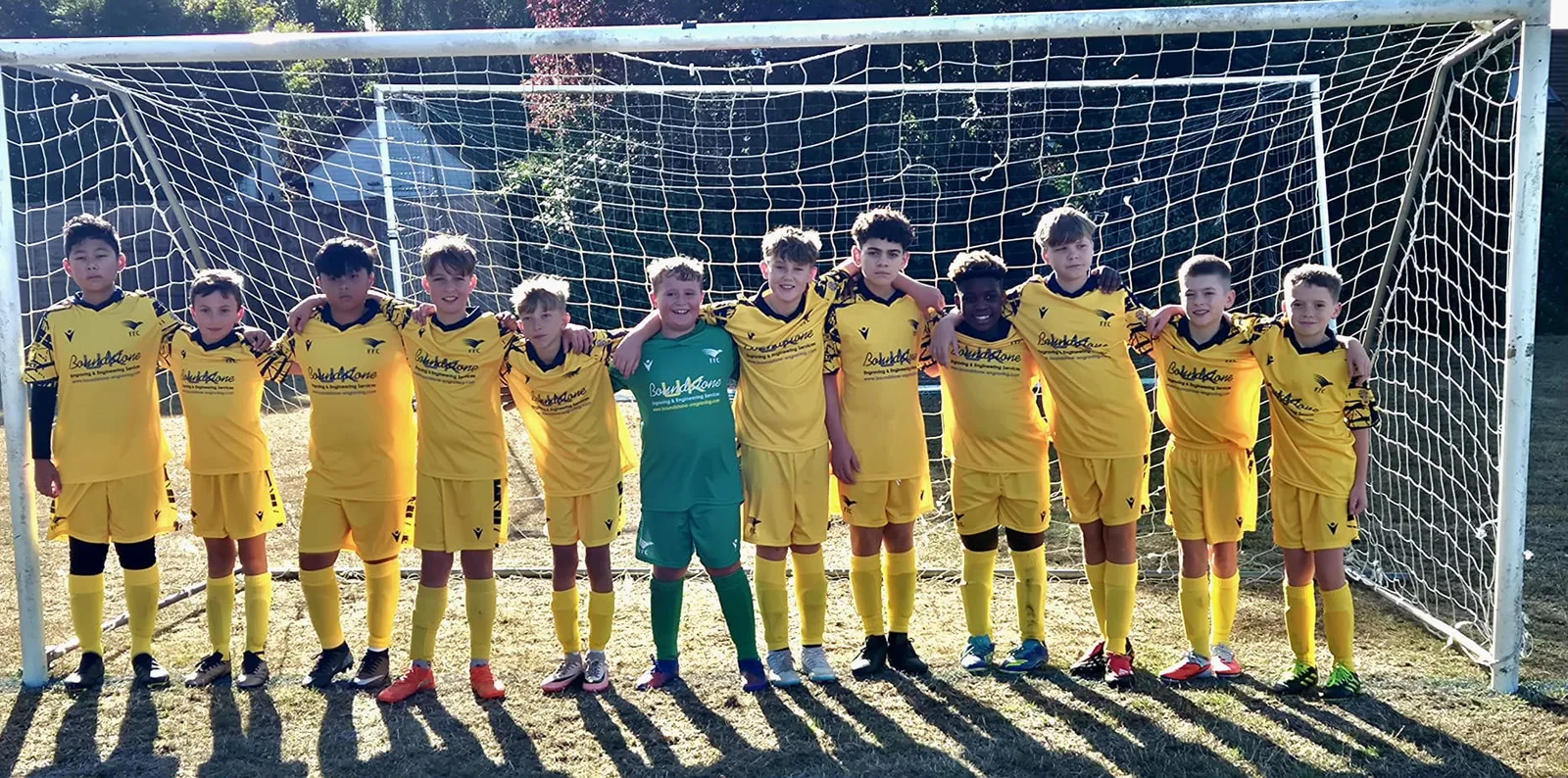 The Under 12 Maroons squad lined up arm in arm across the goalmouth, the goalkeeper in green in the middle, long shadows on a dry pitch