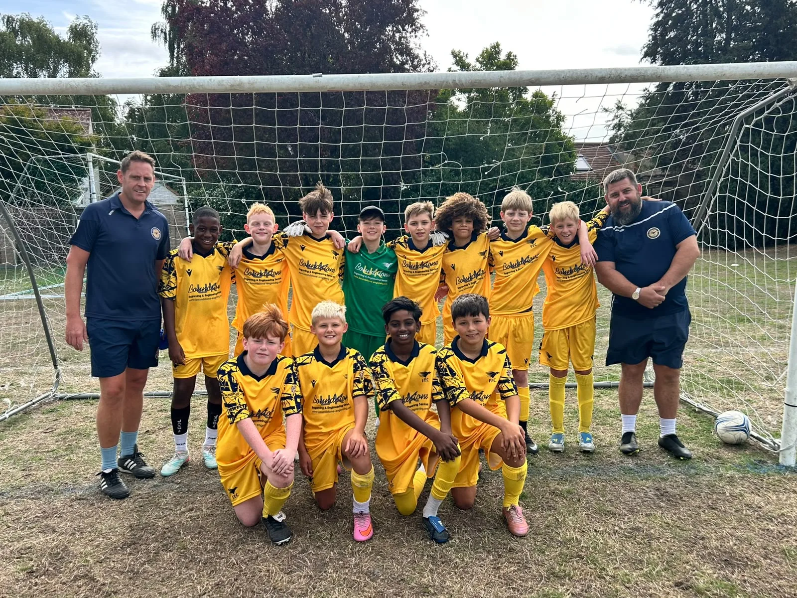 The Under 12 Yellows squad and their two coaches in front of the goal, eight standing and four kneeling in front, the goalkeeper in green