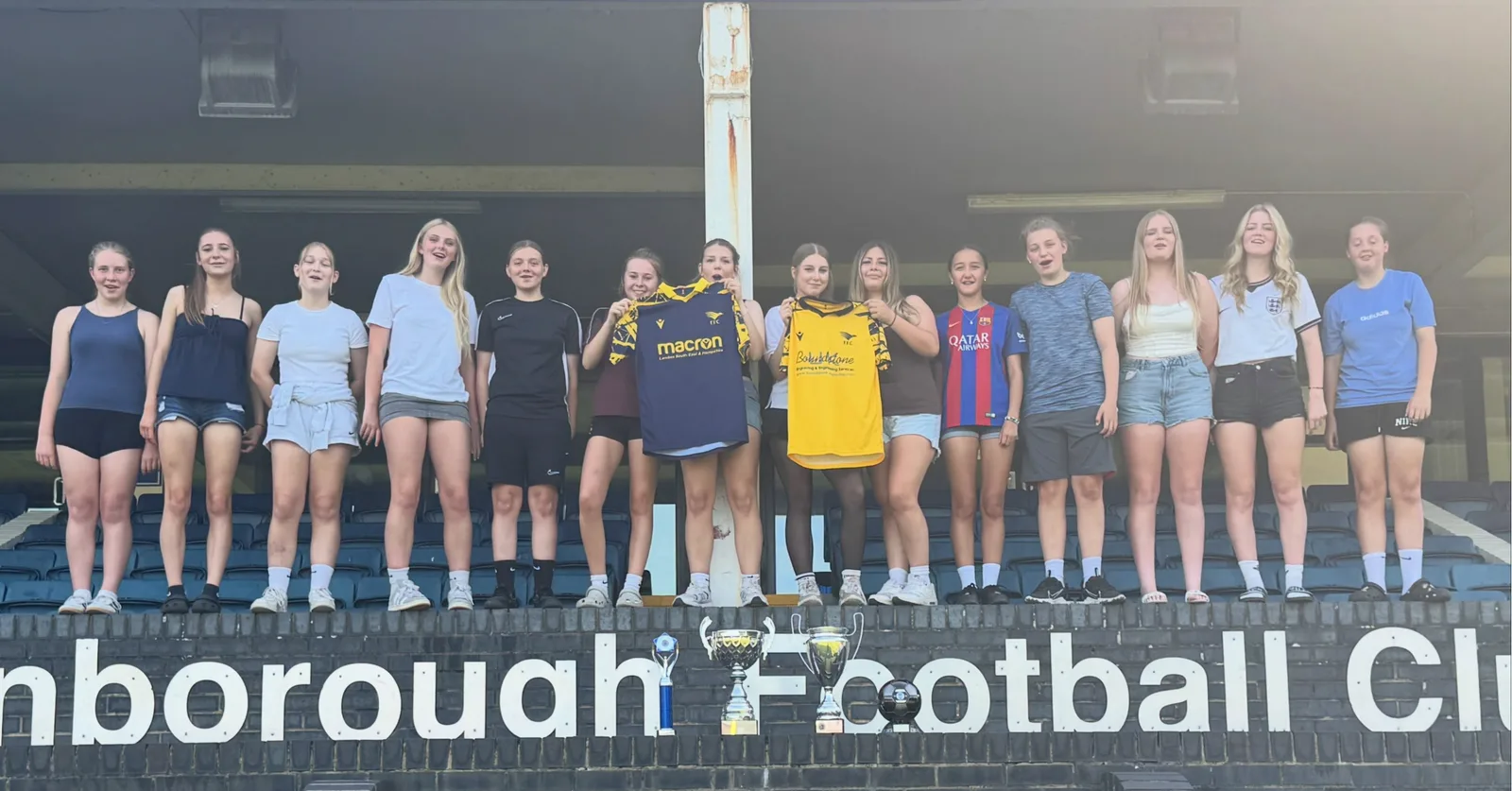 The Under 16 Girls lined up along the front of the stand at Farnborough FC, holding up the home and away shirts, with three trophies and a ball on the wall below
